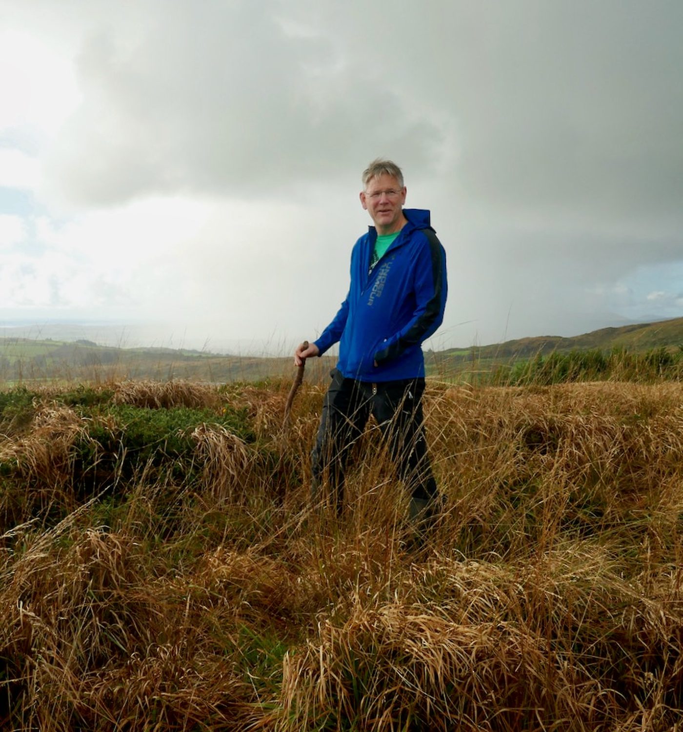 Oliver standing on the mountain in a blue jacket with a walking stick, with rolling land and hills behind him