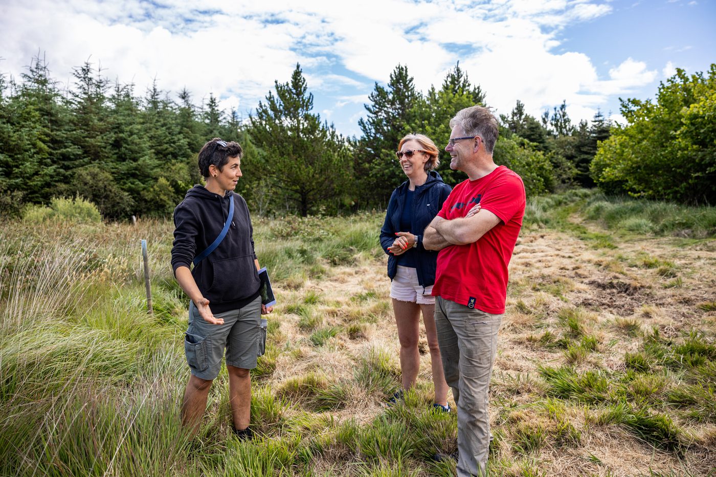 Three people standing in a forest clearing in conversation, with pine trees and moorland grass behind them