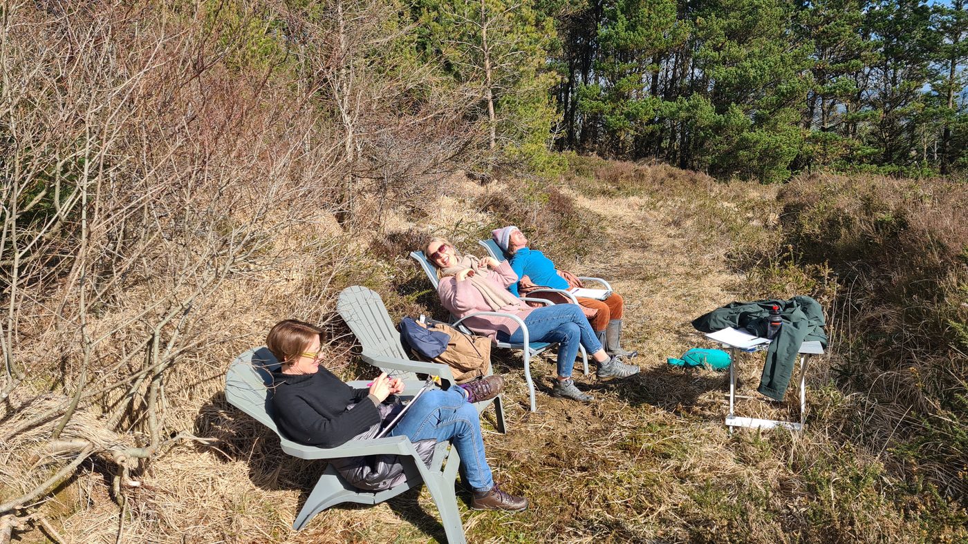 Three people sitting in folding chairs outdoors, laughing with papers in hand