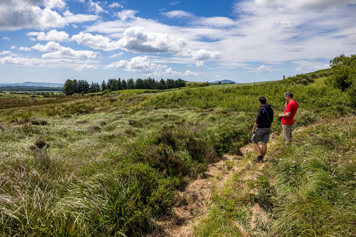Two people standing on a grass trail on a hillside, looking towards fields and the sea
