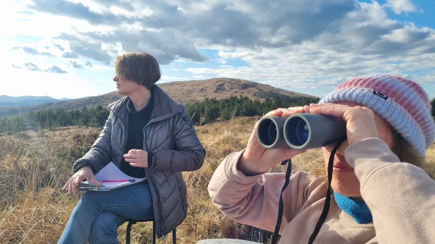 Two women on the land, one writing in a notebook and one looking through binoculars, with a mountain behind them