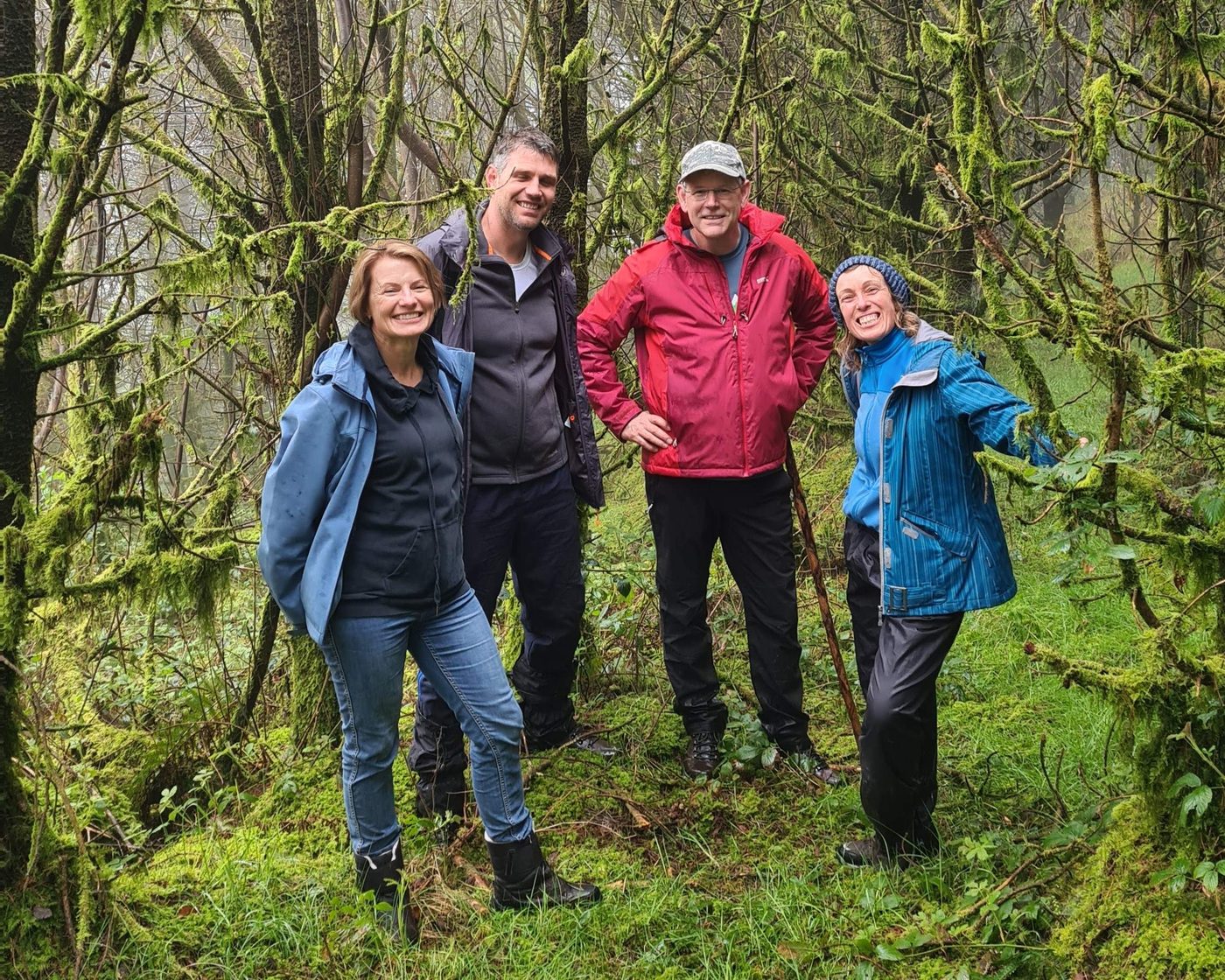 Four people standing together in a mossy, misty forest, smiling at the camera